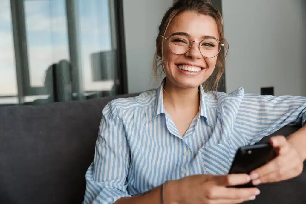 A cheerful woman in glasses and a striped shirt, holding a phone, interested in Teeth Whitening at Laughlin Dental